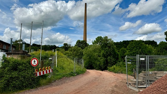 Eine Baustelle umgeben von Bäumen und Sträuchern. Im Hintergrund ragt ein Schornstein ins Bild.