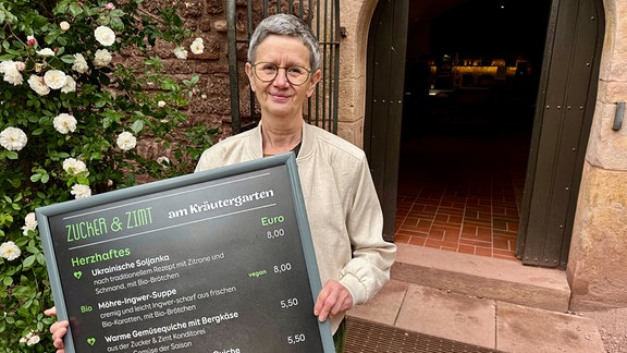 Eine Frau hält vor einer geöffneten Tür eine großes Speisekarten-Schild in der Hand.