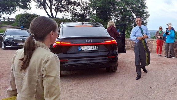 Ein Mann im Anzug läuft neben einem Auto auf einem Platz. Drumherum stehen Zuschauer.