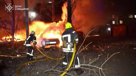 Zwei Kinder unter den Toten nach russischem Raktenangriff in Sumy, zwei Feuerwehrmänner löschen ein brennendes Auto.