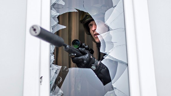 Scharfschütze schaut aus einem Fenster. Das Gewehr trägt einen Schalldämpfer.