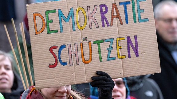 Demonstrierende halten ein Plakat „Demokratie schützen“ hoch. 