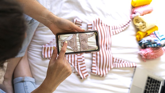 Eine Frau sitzt auf dem Bett und macht mit dem Smartphone ein Foto von einem Bikini