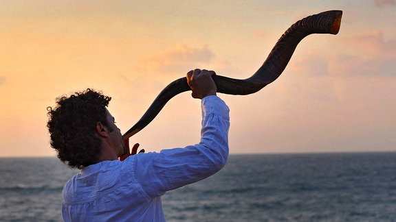 Ein Mann bläßt am Meer stehend in ein mehrfach geschwungenes Horn.