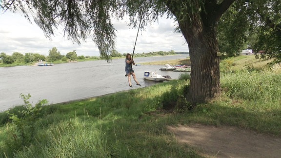 Kind schaukelt an einem Baum, im Hintergrund ein Fluss.