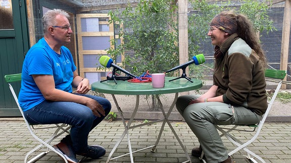 Ein Mann und eine Frau sitzen sich in einem Tiergehege an einem Tisch mit Mikrofonen gegenüber.