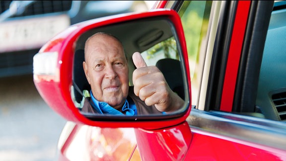 Ein älterer Mann sitzt im Auto und spieglt sich im Seitenspiegel.