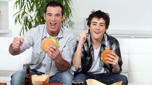 Vater und Sohn sitzen auf dem Sofa, essen Burgen und freuen sich.