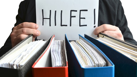 Ein Zettel mit der Aufschrift 'Hilfe' wird hinter Aktenordnern gehalten.