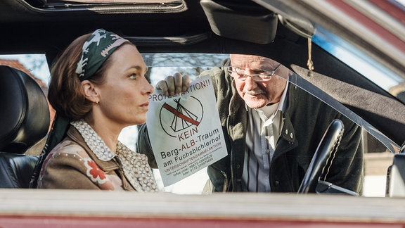 Sophie (Aglaia Szyszkowitz) sitzt in einem Pkw und blickt starr nach vor. Barthl (Friedrich von Thun) blickt durch ein Seitenfenster zu ihr. Er hält ein Flugblatt "Kein Berg-Alb-Traum" in das Fahrzeug.