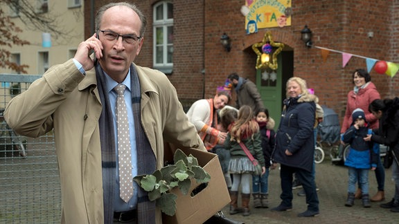 Stenzel (Herbert Knaup) hat dem Kindergarten einen "Sonderkredit" ermöglicht und wird von der Leiterin Rubin (Regine Hentschel, hinten Mitte) zur Eröffnung eingeladen. 