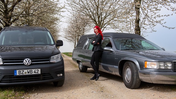 EIne Frau mit knallroten Haaren lehnt an einem Bestattungsauto.