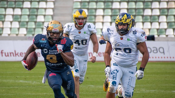 Szene aus einem Footballspiel: Ein Spieler in blau läuft mit Ball zweien in weiß davon.