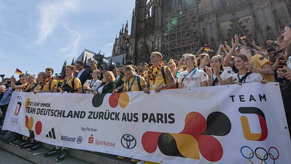 Olympia-Fans auf dem Domplatz in Köln halten ein Willkommensplakat.