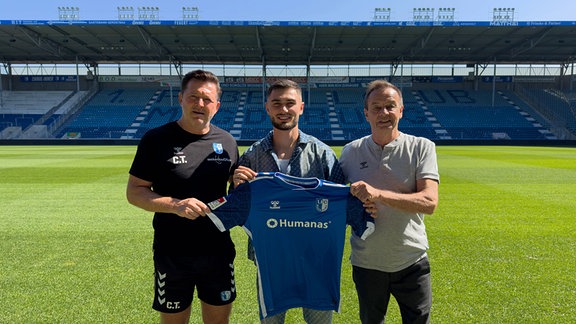 FCM-Neuzugang Samuel Loric mit einem Trikot der Magdeburger umrahmt von Trainer Christian Titz und Geschäftsführer Sport Otmar Schork auf dem Rasen des Stadions der Elbestädter.