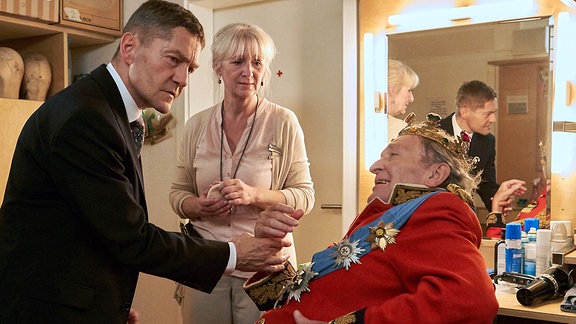 Dr. Rolf Kaminski (Udo Schenk, li.) wird bei einem Theaterbesuch in die Garderobe von Joseph Bernau (Michael Mendl, re. ) gerufen. Während er von Maskenbildnerin Rita (Kerstin Aedtner, mi.) geschminkt wurde, hatte er plötzlich starke Schmerzen. Um das abzuklären, muss die Vorstellung ausfallen und Kaminski nimmt Bernau mit in die Sachsenklinik.