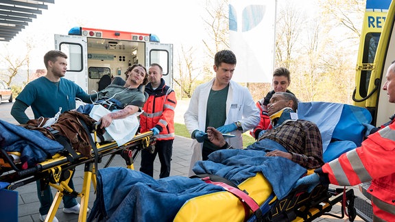 Mikko Rantala (Luan Gummich, l.) und Ben Ahlbeck (Philipp Danne, M.) übernehmen die Patienten von den Rettungsanitäter:innen (Kompars:innen).