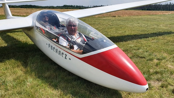 Zwei Personen in einem kleinen, rot-weißen Segelflugzeug auf dem Boden.