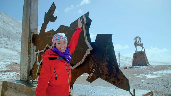 Eine Frau steht vor einer Stahlplatte, der Karte Tadschikistans, im Hintergrund ist ein Widder-Denkmal.