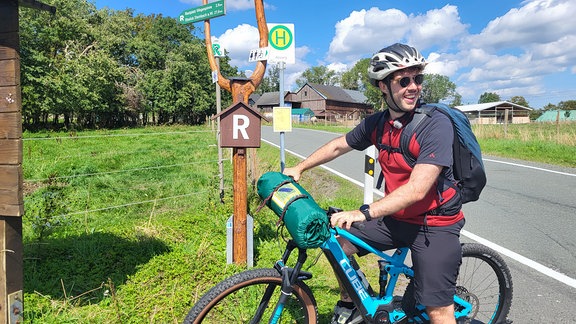 Ein Mann mit Sonnenbrille steht mit seinm Fahrrad an einen Rennsteig-Wegweiser.