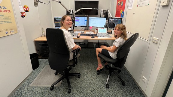 Moderatorin Tiffany Zimmermann und Maya aus Halle sitzen vor dem Mischpult im Radiostudio. Im Hintergrund ist der Schreibtisch mit vielen Knöpfen und Rgelern, Mikros und Bildschirmen zu sehen. 