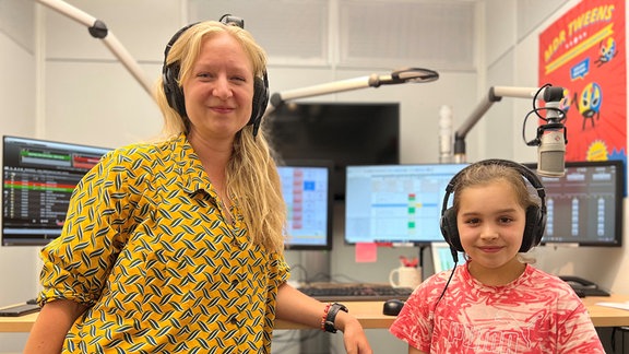 Moderatorin Vivi und Studiogast Paulina stehen nebeneinander im MDR Tweens Studio in Halle. Sie schauen beiden in die Kamera, hinter ihnen sind ein Mischpult, Bildschirme und viele Knöpfe zu erkennen. 