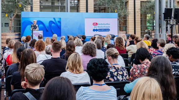 Eine Frau steht auf einer Bühne vor einem blauem Bild. Vor sitzen viele Menschen in Reihen.