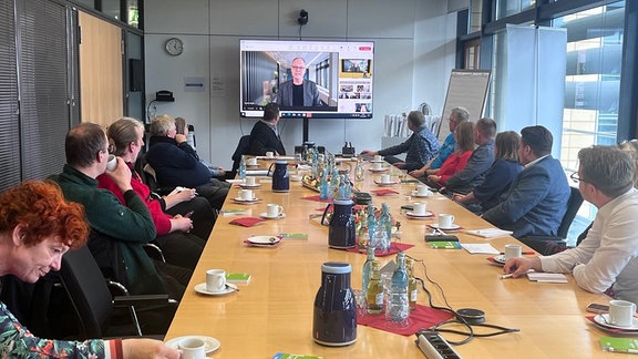 Menschen an einem Tisch sitzend schauen auf Videoleinwand