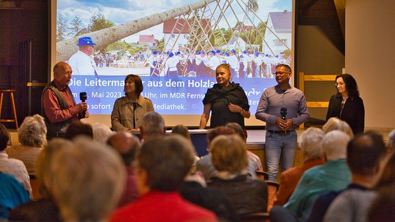 Drei Frauen und zwei Männer stehen auf einer Bühne vor Publikum. Im Hintergrund ist ein Bild auf einer Leinwand zu sehen.