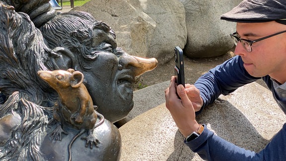 Mann hockt auf Felsen und fotografiert Hexen-Figur.