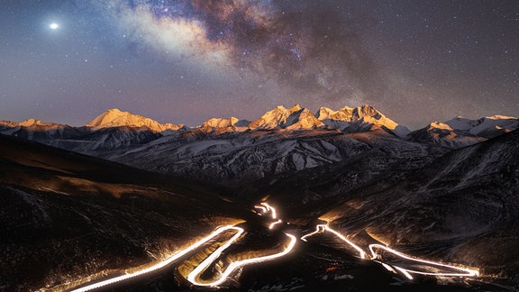 Kategorie Mensch und Weltall: "Der Sternenhimmel über der höchstgelegenen Nationalstraße der Welt" (© Yang Sutie) - Der beleuchtete National Highway 219, die höchstgelegene Nationalstraße der Welt, schlängelt sich durch den Vordergrund und spiegelt fast das majestätische Bild der Milchstraße darüber wider. Die beiden sind durch den Kula Kangri getrennt, einen Berg in der Präfektur Shannan in Tibet.