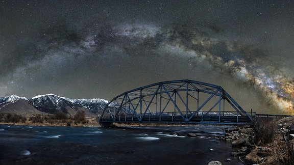 Kategorie: Himmelslandschaften  Überquerung des Madison © Jake Mosher  Die Milchstraße wölbt sich über der Three Dollar Bridge in Montana im April 2021. Der Fotograf fuhr 100 Meilen weit, um den Kern der Milchstraße ein paar Stunden vor Tagesanbruch über den Horizont steigen zu sehen.   Aufgenommen mit Nikon D850 Kamera, 35mm f/4, ISO 1600/640, Mehrfachbelichtungen zwischen 110 und 180 Sekunden  Standort: Madison River, Montana, USA 