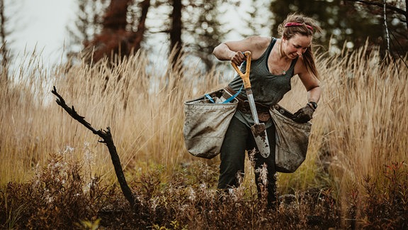 Frau mit Stirnband, Top und Handschuhen, Schaufel und zwei Säcken in einem Wald, um ein Pflanzloch zu graben