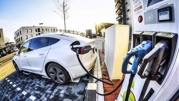 Weitwinklige Aufnahme mit Fischaugen-Verzerrung: Ladesäule mit verschiedenen Steckern für Elektroautos, ähnlich wie eine Zapfsäule, und ein Tesla-Elektroauto, das mit der Säule durch ein Kabel verbunden ist.