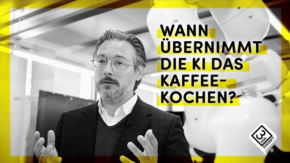 Mann mit silberner, drahtiger Brille mit gebogenem Steg und Bart blickt in Kamera, Vordergrund unscharf Roboterarm, Hintergrund unscharf Werkstatt-Aufbau. Erklärende Gestik. Text: "Wann übernimmt die KI das Kaffeekochen?"