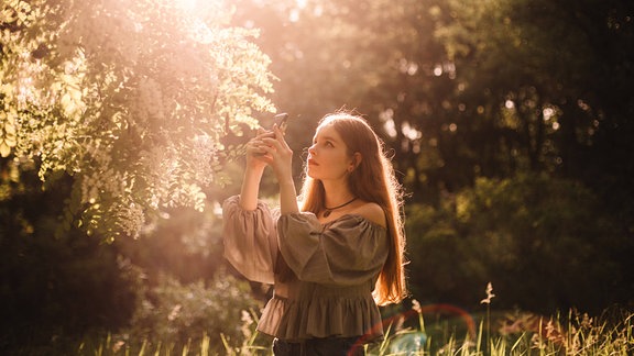 Jugendliche oder junge Frau steht konzentriert im sommerlichen Gegenlicht vor Blütenbaum und macht Foto mit Smartphone von Blüten