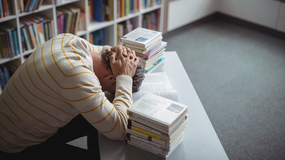 Foto leicht von obene: Lehrer mit Kopf auf Tisch, Hände an Hinterkopf, links und rechts Bücherstapel, dahinter Buchregal