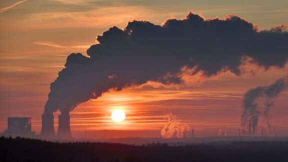 Zwei große Kraftwerkstürme bei Sonnenuntergang, viele Schleierwolken, viel wolkiger Ausstoß aus Türmen, tief-rote Stimmung, untergehende Sonne direkt sichtbar. Weitere kleine Schornsteine und kastenförmiger Kraftwerksbau.