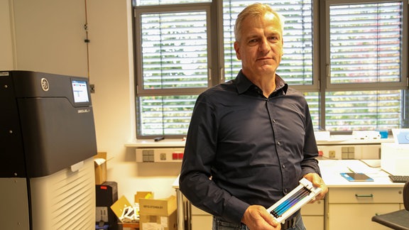 Mann mit grauen Haaren und schwarzem Hemd in einem Labor, links ein druckerähnlicher schwarzer Kasten mit Display, in der Hand ein glänzendes, rechteckiges Utensil. Blick in Kamera. Hintergrund: Labortische, Fenster mit Jalousie.