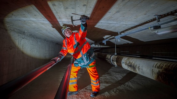 Weitwinkelaufnahme: Person mit Helm und organgefarbenen Signalanzug in Unterflurbühne einer Autobahnbrücke beklopft mit Hammer Beton