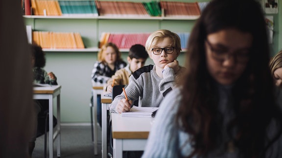 Junge traurig-nachdenklich auf Hand aufgestützt im Klassenzimmer, im Vordergrund unscharf und dunkel belichtet Mädchen, im Hintegrund unscharf weitere Schülerinnen und Schüler und Buchregal.