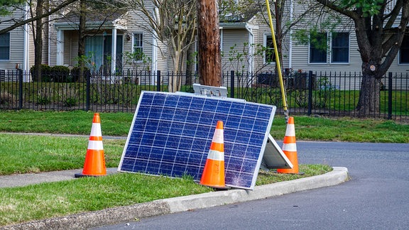 Angeknackstes Solar-Modul steht an einen Mast gelehnt an Straße mit Warnkegeln, im Hintergrund Haus