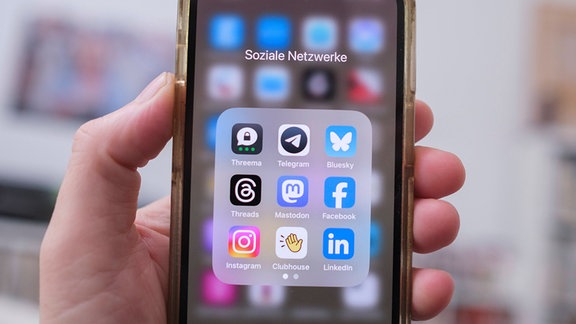 Hand hält frontal zur Kamera iPhone mit geöffnetem App-Ordner Soziale Netzwerke, darin Apps unter anderem von Mastodon, Instagram, Bluesky und Facebook