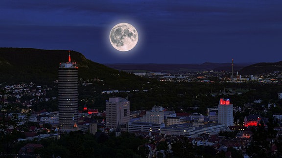 Fotomontage: Skyline von Jena mit mehreren Hochhäusern bei Nacht, einige rote Lichter, großer Vollmond am Himmel im Hintergrund