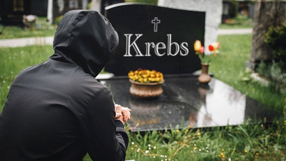 Mann trauert auf einem Friedhof vor einem Grabstein. Grab mit Aufschrift: Krebs, Symbolbild 