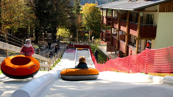 Blick von oeben auf eine Rodelbahn aus Kunstfaser. Ein Kind fährt in einem Reifen den Hang hinunter. 