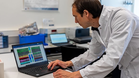 Junger Forscher in weißem Kittel beugt sich über einen Laptop, darauf zu sehen verschiedene bunte Grafiken.