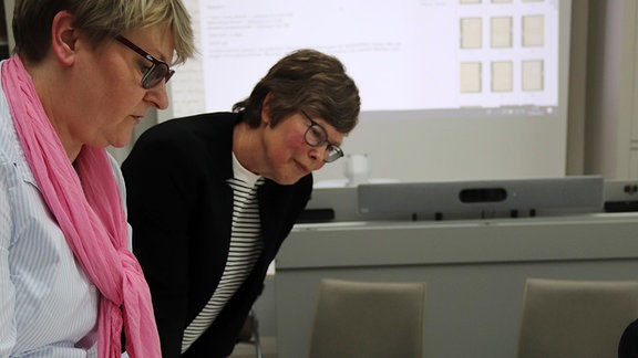 Zwei Frauen beugen sich über einen Tisch, auf dem eine aufgeschlagene Mappe mit bedrucken Seiten liegt. Im Hintergrund ist eine Leinwand zu sehen, auf die eine Präsentation mit Manuskriptseiten projiziert wird.